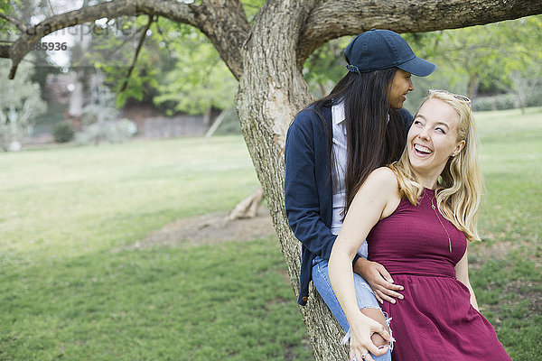 Lächelndes junges lesbisches Paar sitzt auf einem Baumstamm im Park.