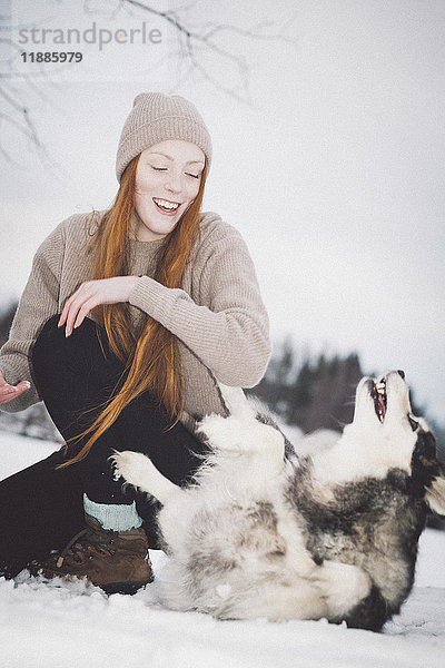 Fröhliche junge Frau spielt mit Siberian Husky auf schneebedecktem Feld