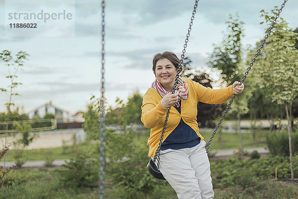 Lächelnde Frau spielt auf Schaukel auf dem Spielplatz gegen den Himmel