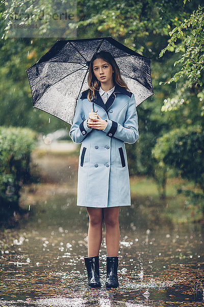 Porträt eines jungen Mädchens mit Regenschirm  das auf dem Weg zwischen Bäumen steht.
