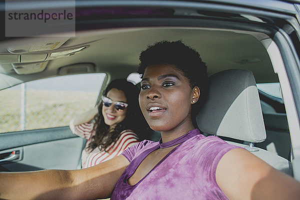 Junge Frau sitzt mit Freundin im Auto und schaut weg  während sie an einem sonnigen Tag im Auto sitzt.