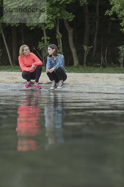 Volle Länge von Mutter und Tochter  die sich beim Hocken am Seeufer im Park unterhalten.
