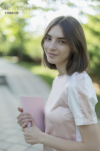 Porträt einer lächelnden jungen Frau  die im Park steht und ein digitales Tablett hält.