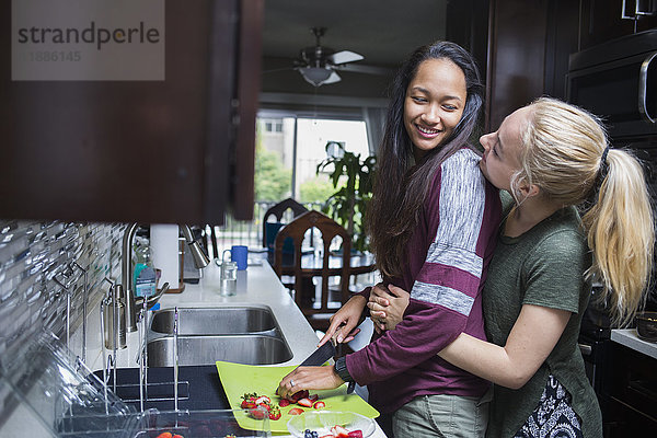 Happy blonde Frau umarmt Freundin Schneiden Erdbeeren in der Küche zu Hause