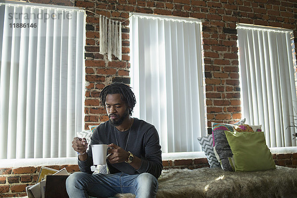 Mann auf dem Bett sitzend mit Kaffeetasse an den Fenstern zu Hause