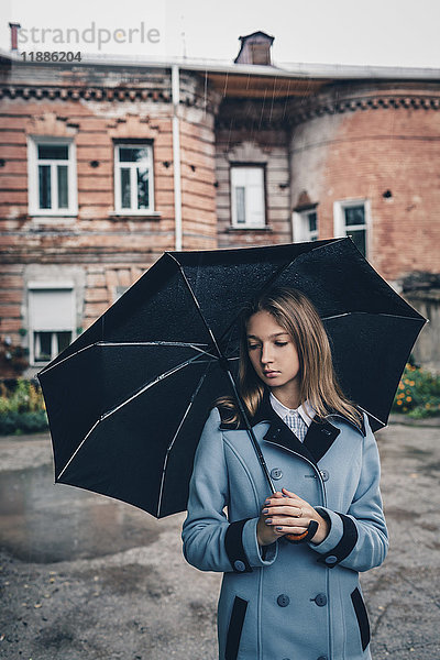 Nachdenkliches Teenagermädchen mit Regenschirm gegen das Gebäude stehend