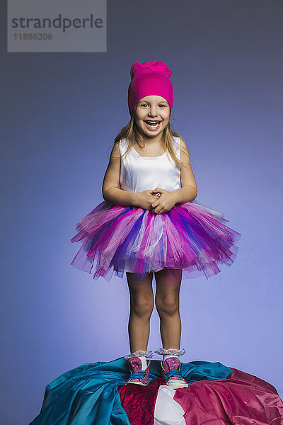Porträt eines fröhlichen Mädchens mit Tüllrock und Strickmütze auf Stoffhaufen stehend gegen lila b