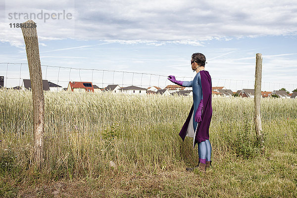 Mann im Superheldenkostüm steht am Zaun auf grasbewachsenem Feld gegen den Himmel.