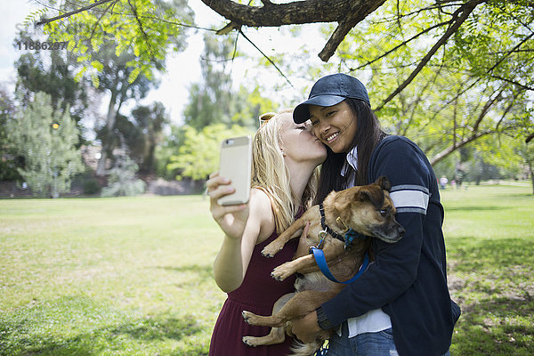 Frau  die sich küsst  während sie Selfie durchs Handy mit Hund im Park nimmt.