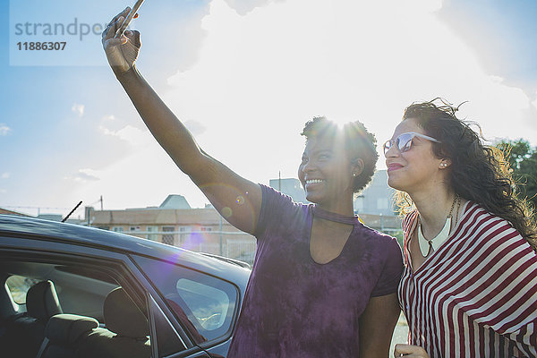 Hinterleuchtete  fröhliche junge Frau nimmt Selfie mit Freundin mit dem Auto gegen den Himmel.