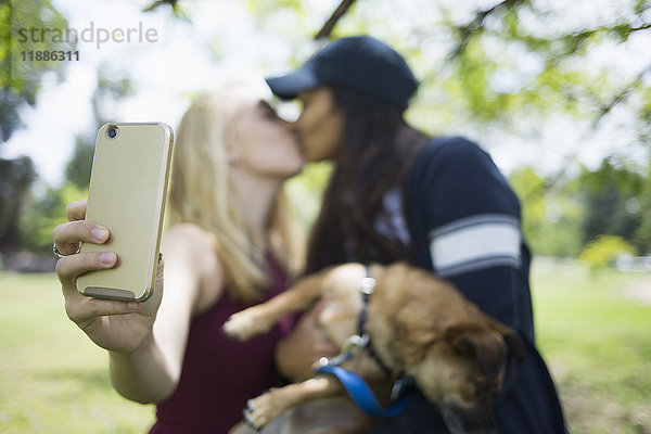 Frauen küssen sich  während sie Selfie durch Smartphone mit Hund im Park nehmen.