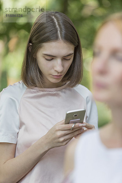 Tochter mit Smartphone  während die Mutter im Park im Vordergrund steht.
