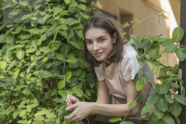 Porträt einer lächelnden jungen Frau  die sich inmitten von Pflanzen auf das Geländer stützt.