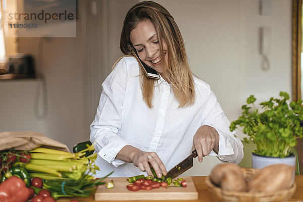 Lächelnde Frau am Telefon beim Gemüsehacken in der Küche