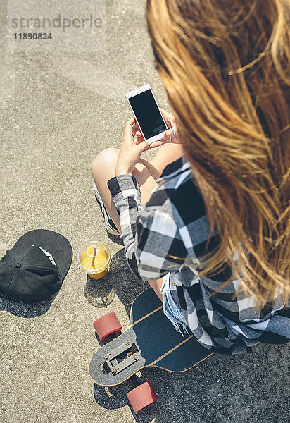 Rückansicht einer jungen Frau mit langen Haaren  die auf dem Longboard sitzt und auf das Smartphone schaut.