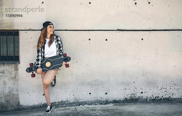 Porträt einer glücklichen jungen Frau mit Longboard vor der Betonwand stehend