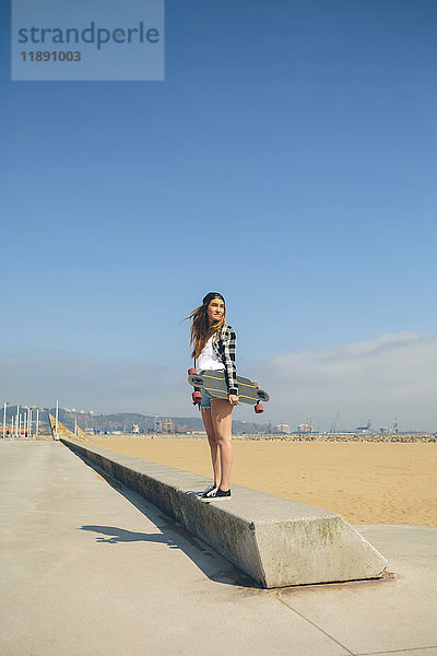 Junge Frau mit Longboard an einer Wand an der Strandpromenade stehend
