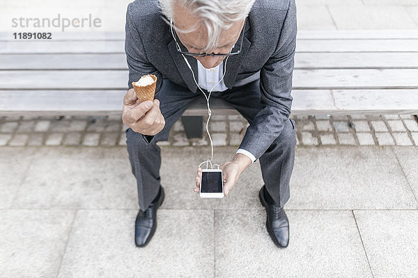 Senior Geschäftsmann mit Eistüte auf der Bank sitzend mit Blick auf das Handy