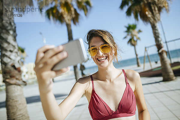 Lachende junge Frau mit gelber Sonnenbrille  die einen Selfie auf der Strandpromenade trägt.