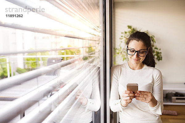 Lächelnde Frau steht am Fenster zu Hause mit dem Smartphone