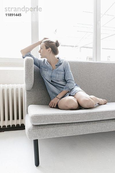 Frau sitzt auf der Couch in einem Loft und schaut aus dem Fenster.