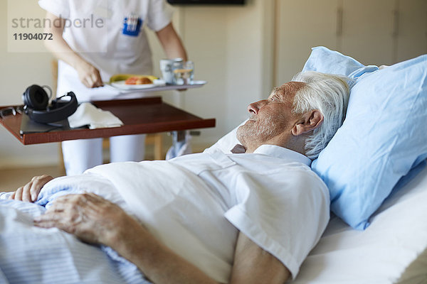 Älterer Mann schaut auf eine Krankenschwester  die das Frühstück serviert  während sie auf dem Krankenhausbett liegt.
