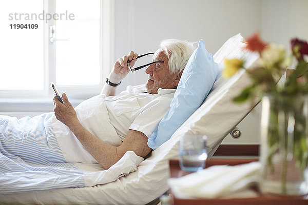 Seitenansicht des älteren Mannes mit Brille bei der Benutzung des Smartphones in der Krankenstation