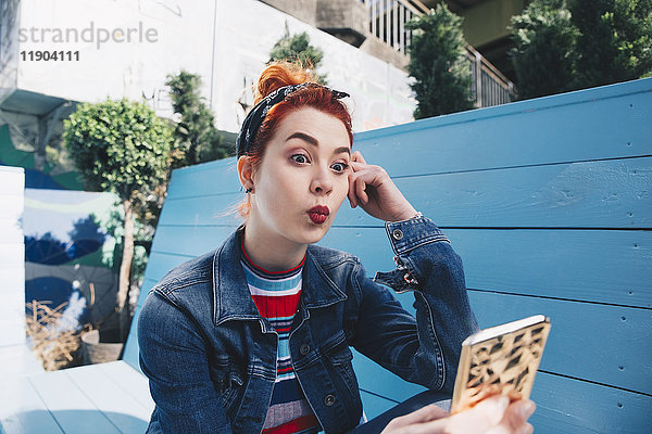 Rothaarige junge Frau nimmt Selfie mit Handy  während sie auf der Bank sitzt.