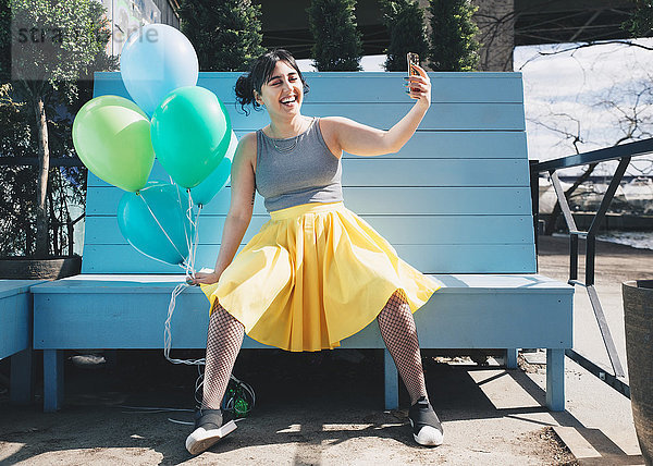 Fröhliche junge Frau nimmt Selfie mit Luftballons mit auf die Bank