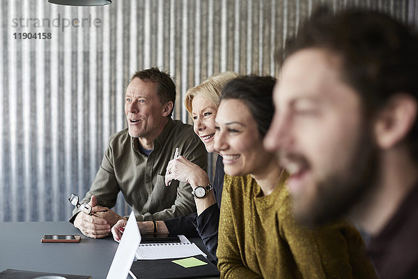 Lächelnde  kreative Geschäftskollegen sitzen am Konferenztisch und schauen weg in den Sitzungssaal.