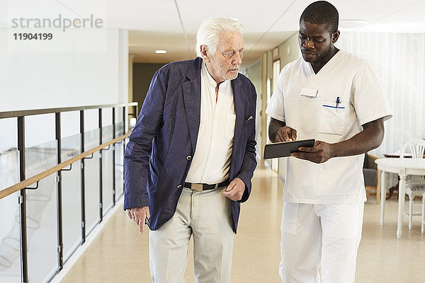 Krankenpfleger mit digitalem Tablett und Senior-Patient im Krankenhausflur
