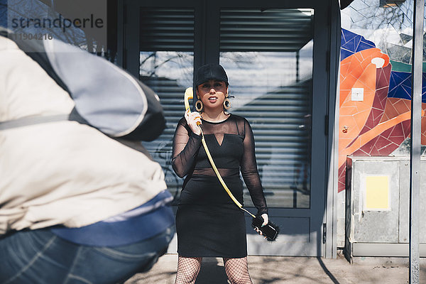 Porträt einer jungen Frau  die einen mit dem Handy verbundenen Telefonhörer hält  während sie gegen das Gebäude steht.
