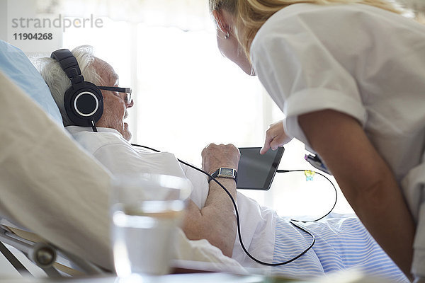 Seitenansicht der Krankenschwester  die den älteren Mann bei der Benutzung des digitalen Tabletts auf dem Krankenhausbett unterstützt.