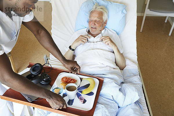 Hochwinkelansicht des Krankenpflegers  der das Frühstück für den älteren Mann auf der Krankenstation serviert.