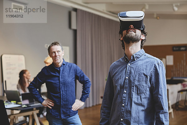 Der reife Geschäftsmann steht mit den Händen auf der Hüfte  während er den männlichen Kollegen mit Hilfe des Virtual-Reality-Simulators in kreativen
