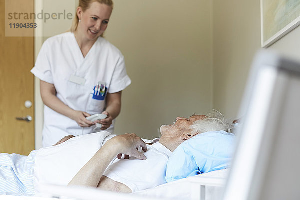 Lächelnde Krankenschwester  die das Bett eines älteren Mannes in der Krankenstation anpasst.