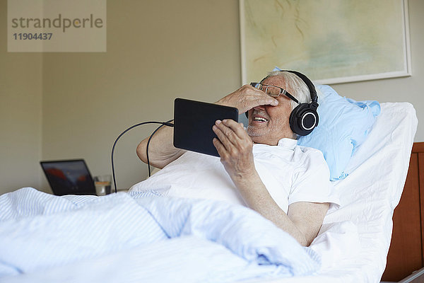 Senior Mann reibt sich die Augen  während er ein digitales Tablett auf dem Bett in der Krankenstation benutzt.