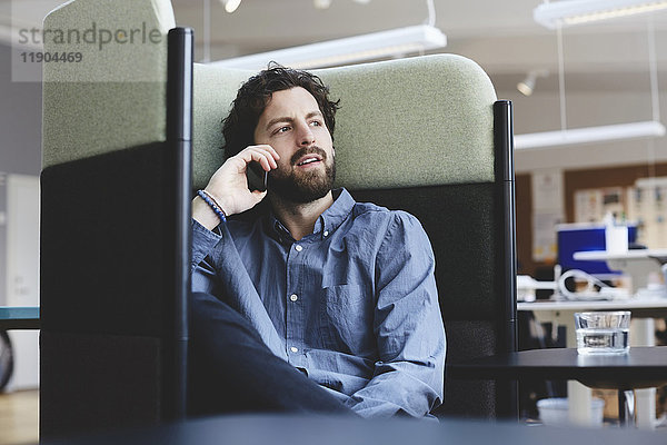 Mittlerer Erwachsener  professioneller Mann  der über Smartphone spricht  während er im Kreativbüro auf dem Stuhl sitzt.