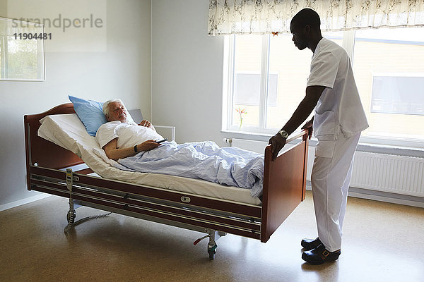 Krankenpfleger  der das Bett anpasst  während ein älterer Mann auf der Krankenstation fernsieht.