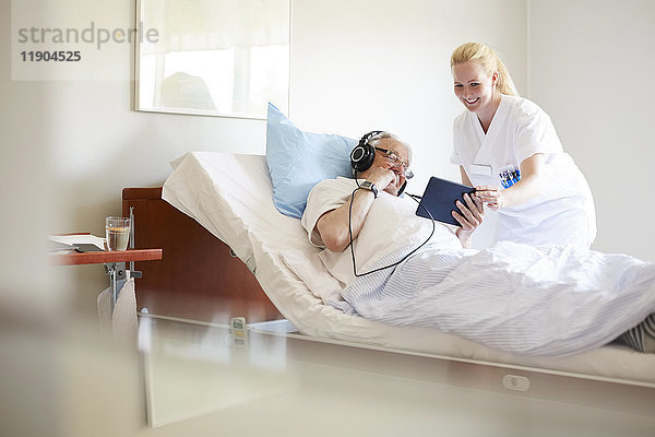 Fröhliche Krankenschwester hilft dem älteren Mann bei der Verwendung von digitalen Tabletten auf dem Krankenhausbett