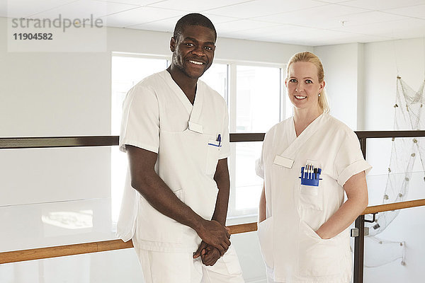 Porträt von glücklichen Krankenschwestern und Krankenpflegern  die am Geländer des Krankenhausflurs stehen.