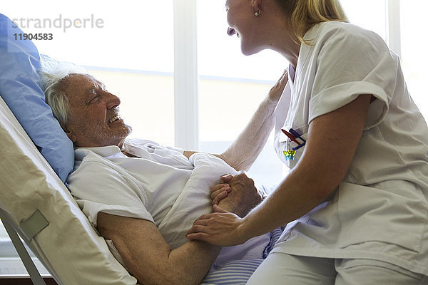 Seitenansicht eines glücklichen älteren Mannes im Gespräch mit einer Krankenschwester im Krankenhausbett