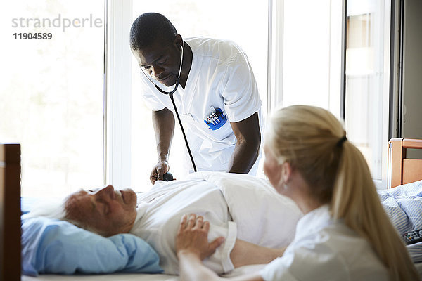 Krankenschwester tröstet älteren Mann  während männlicher Kollege mit Stethoskop auf der Krankenstation untersucht.