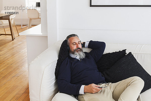 Kaukasischer Mann auf dem Sofa  der mit Kopfhörern Handy hört