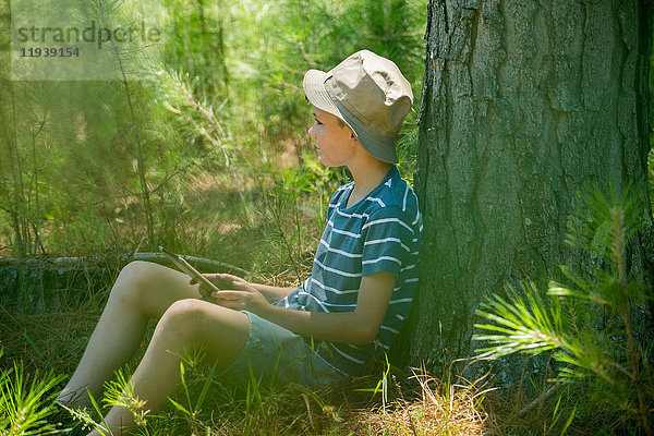 Junge lehnt an Baumstamm mit digitalem Tablett in der Hand