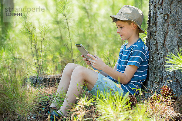 Junge lehnt sich an den Baumstamm  mit digitalem Tablett