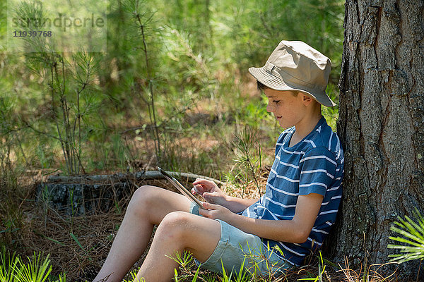 Junge lehnt sich an den Baumstamm  mit digitalem Tablett