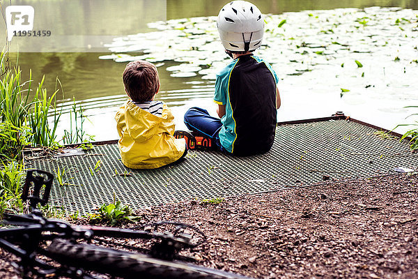 Zwei junge Brüder sitzen am Wasser  Rückansicht