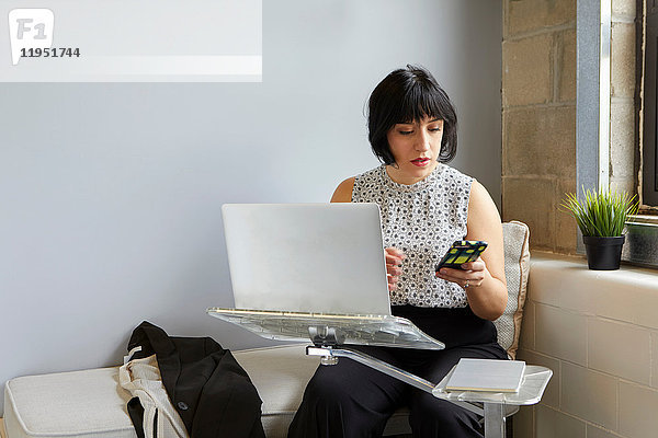 Frau sitzt mit Laptop auf Laptop-Ständer und hält Smartphone