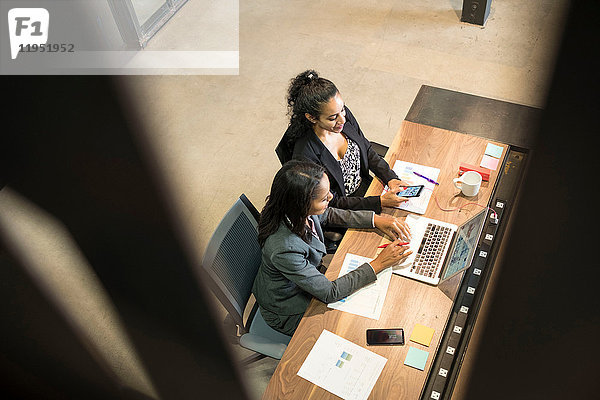 Zwei Geschäftsfrauen sitzen am Schreibtisch  verwenden einen Laptop  schauen auf ein Smartphone  erhöhte Ansicht